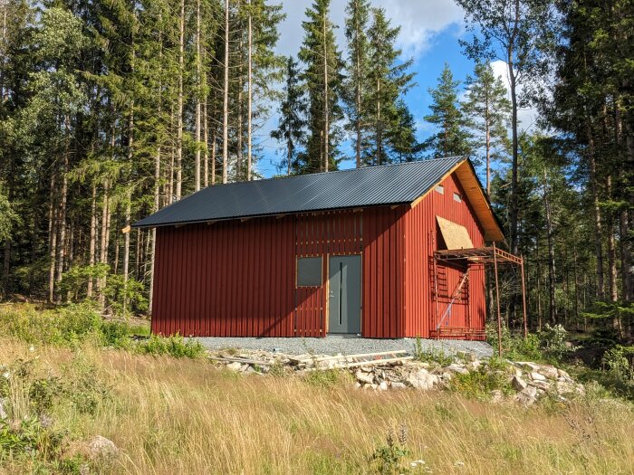 Rött trähus med svart tak omgivet av skog och klarblå himmel.