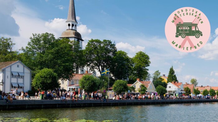 En folksamling vid vattnet, kyrktorn, svenska flaggan, lugn sommardag, Mariefred, evenemangslogotyp.