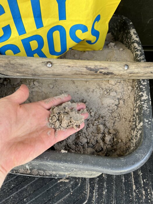 En hand håller sand framför en påse och hjulbåren; kontrast mellan gult och grått.