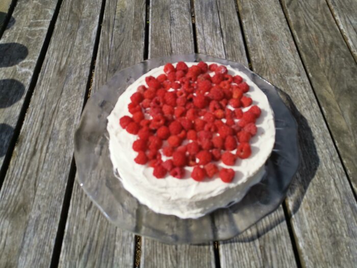 Tårta med grädde och hallon ovanpå, placerad på ett träbord utomhus. Suddig, dagtid.