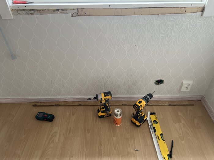 Renovering pågår. Verktyg och material på golv. Oavslutad tapetsering och synliga elledningar. Inredning arbetszon.