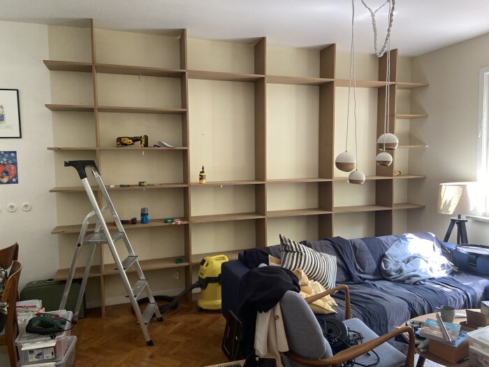 Vardagsrum under omorganisation, stor tom bokhylla, stege, soffa och utspridda föremål.