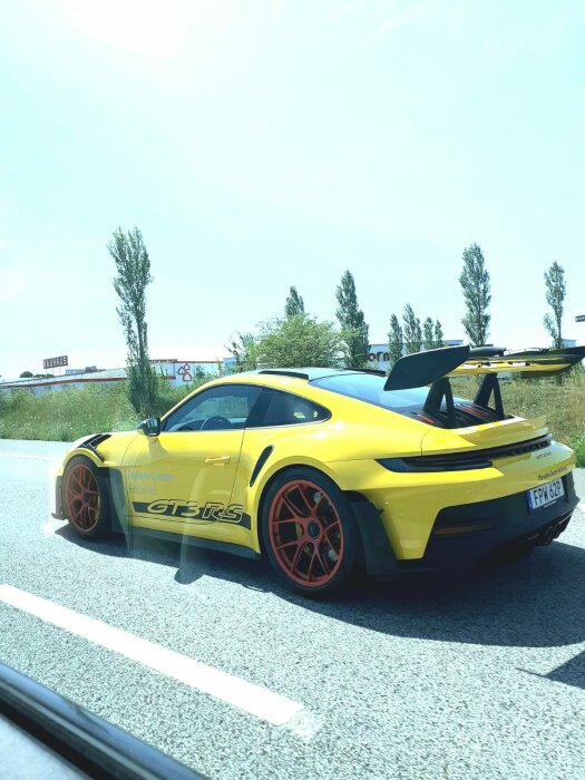 Gul Porsche 911 GT3 RS på motorväg, röda fälgar, bakvinge, klistermärken, dagtid, delvis molnig himmel.