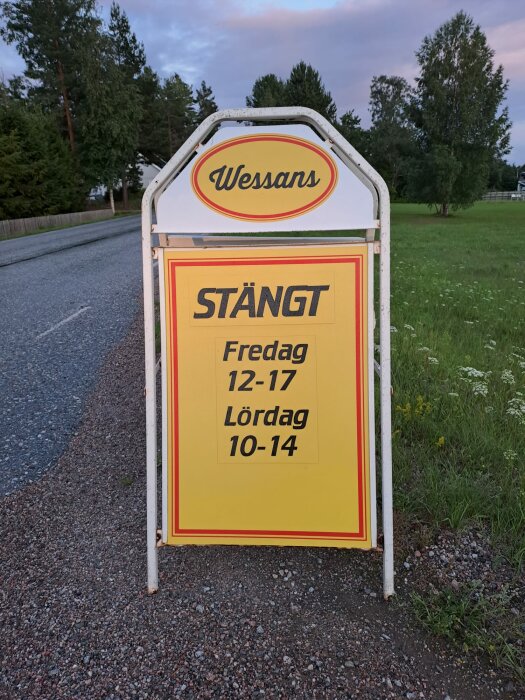 Gatuskylt med öppettider, "Wessans" text, grönt landskap i bakgrunden, skymning eller gryning, asfalterad väg.