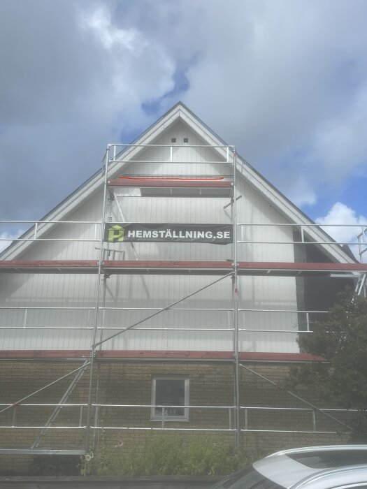 Vit byggnad med byggnadsställning, molnhimmel, reklamskylt "hemställning.se", fönster, grönska, parkerad bil.