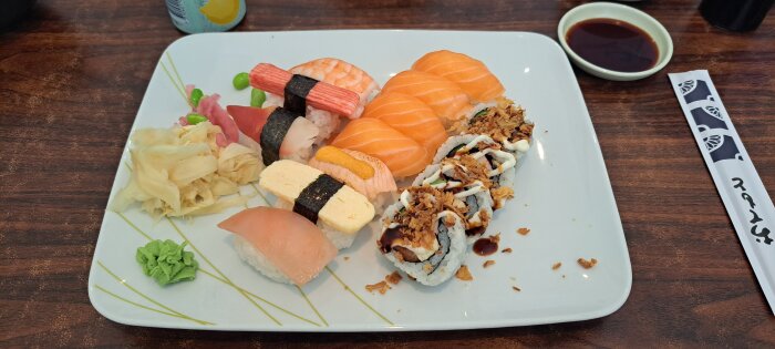 Sushitalrik med nigiri, maki, ingefära, wasabi, sojasås och ätpinnar på träbord.