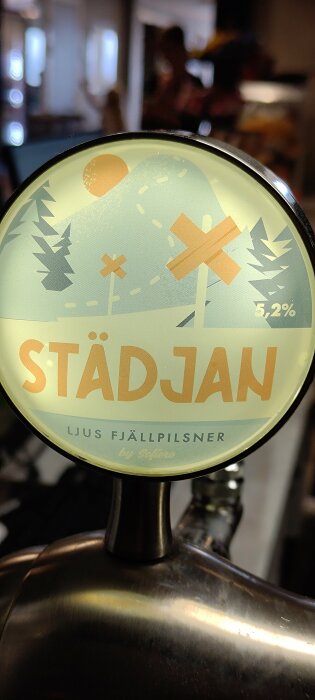 Öl-tapphandtag med etikett "STÄDJAN Ljus Fjällpilsner by Sofiero", 5,2% alkohol, fjällmotiv, inomhus, pubmiljö.