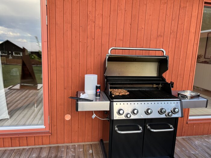 Gasolgrill med mat på, står på träaltan framför röd trävägg och fönster.