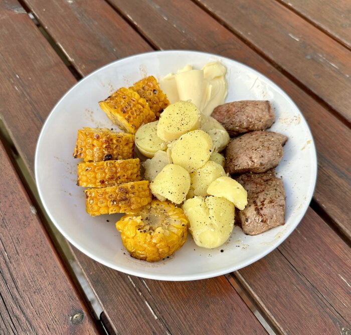 Grillad majs, stekta köttbitar och kokta potatisar med smör på tallrik utomhus.