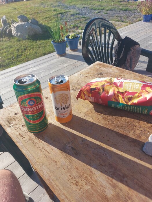 Trädäck med ölburk, ciderburk och chips. Soligt väder, grönområde och trädgårdsmöbler i bakgrunden.