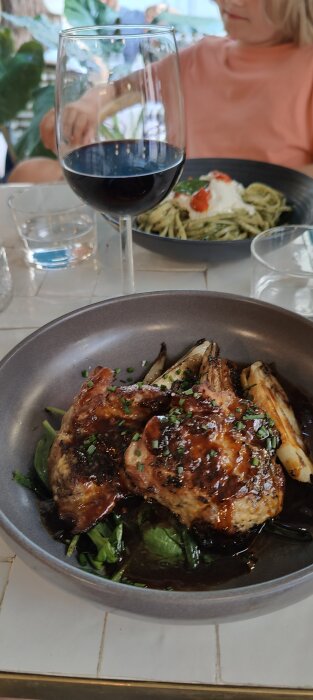 Måltid med grillad kyckling, grönsaker, pasta bakom, vin och vattenglas, person i restaurangmiljö.