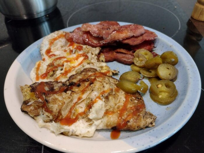 En tallrik med stekt ägg, bacon och jalapeño-skivor toppad med sås. Enkel hemlagad måltid.