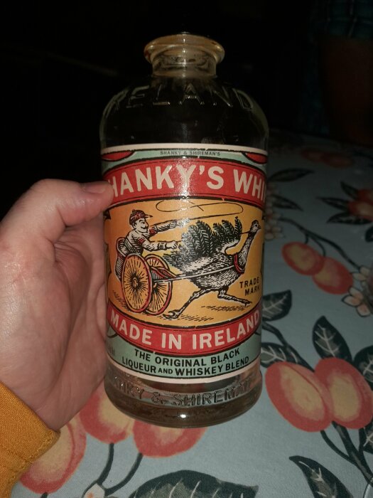 En hand håller en irländsk whiskeyblandflaska, Shankey's, vid ett blommigt bord.