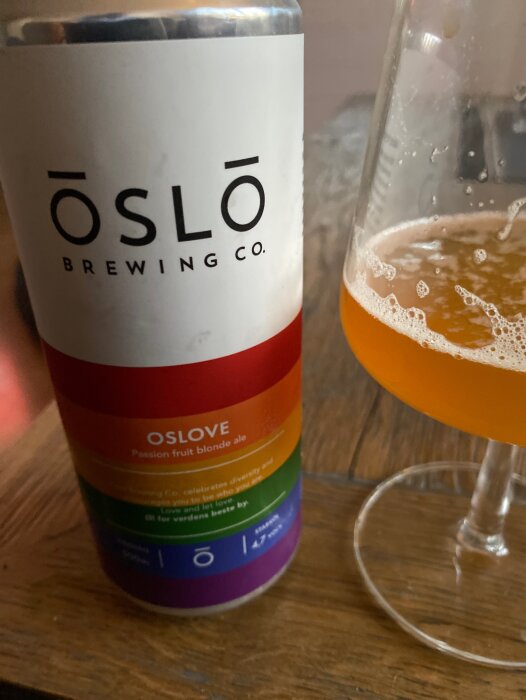 Ölburk från Oslo Brewing Co., regnbågsfärger, "OSLOVE Passion fruit blonde ale", halvfullt ölglas, träyta.