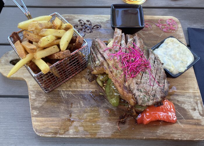 Grillat kött, pommes frites, röd lök, två såser, grillade grönsaker, träbräda, träbord, måltid.