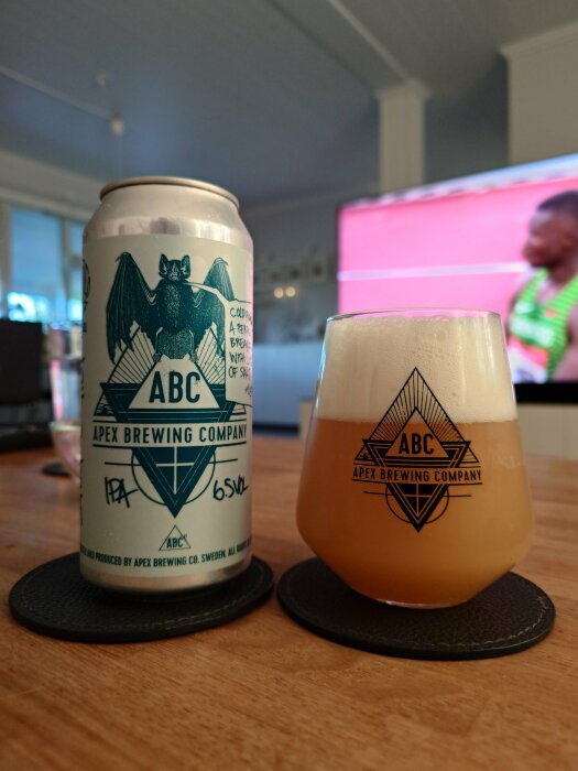 Ölburk och glas med skummande dryck, märkta "Apex Brewing Company", på bord framför suddig TV-skärm.