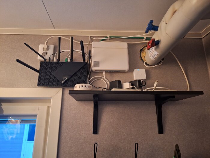 Knökfull hylla med router, switch, sladdar och teknisk utrustning under ett skrivbord.