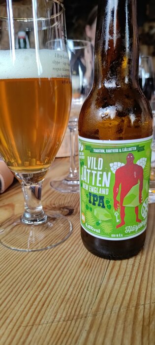 Ölglas med skum vid kanten, ölflaska märkt "Vild Jätten New England IPA". Träbord i bakgrunden.