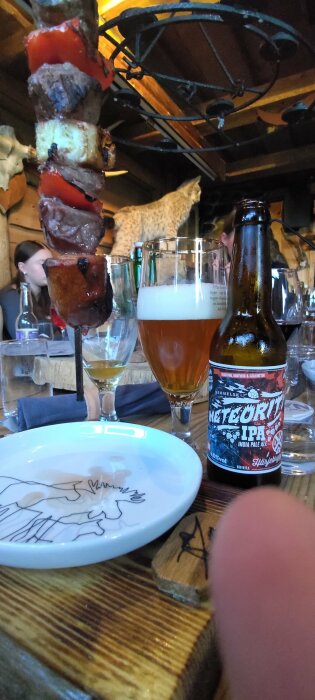 Köttspett över bord, IPA-öl, rustik restaurang, uppstoppad katt i bakgrunden, tom tallrik, sällskapsmiljö.