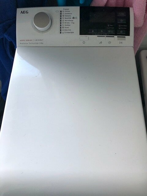Vit topp av en AEG tvättmaskin med reglage och display synlig, placerad nära textilier.