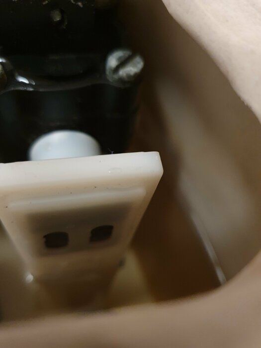 Närbild inuti en toalett, visar delar av flottörventil och mekanism. Otydliga konturer och skuggor i bakgrunden.