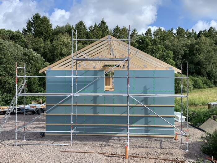 Byggnadsställning framför hus under konstruktion med träspik, blå himmel och träd i bakgrunden.