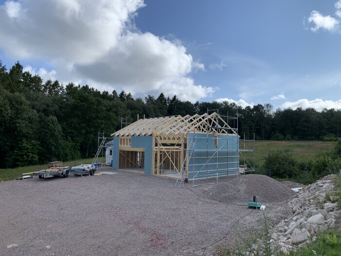 Hus under konstruktion med trästomme, byggnadsställningar, grusplan, skogsbakgrund och molnig himmel.