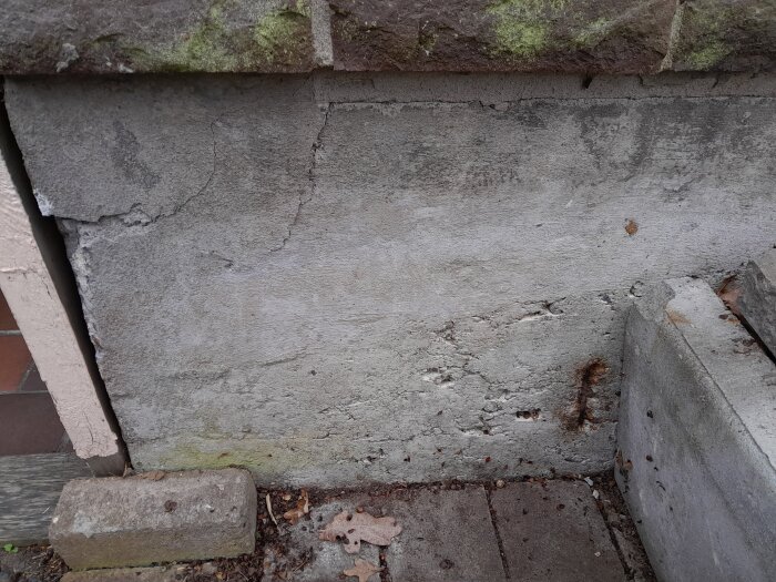 Grå betongvägg med sprickor och slitage, mossa, löv på marken, tegelkant överst.