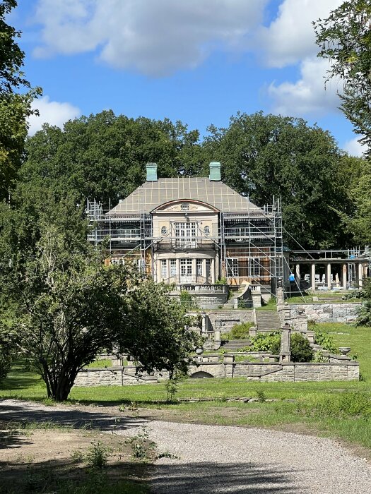 Klassisk byggnad under renovering, omgiven av skog och trädgård, ställning, blå himmel, solljus, moln.