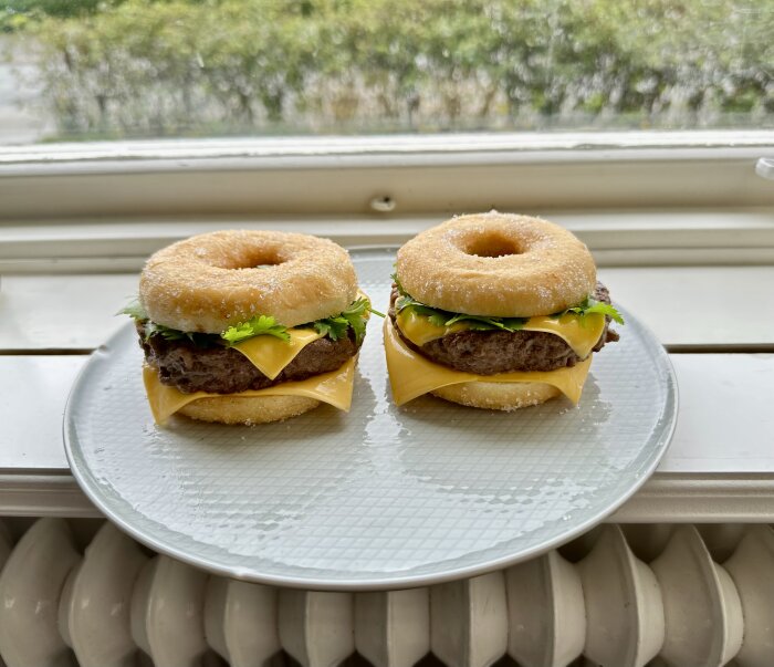 Två hamburgare med munkar istället för bröd, cheddarost, köttpuckar och persilja, framför ett fönster.