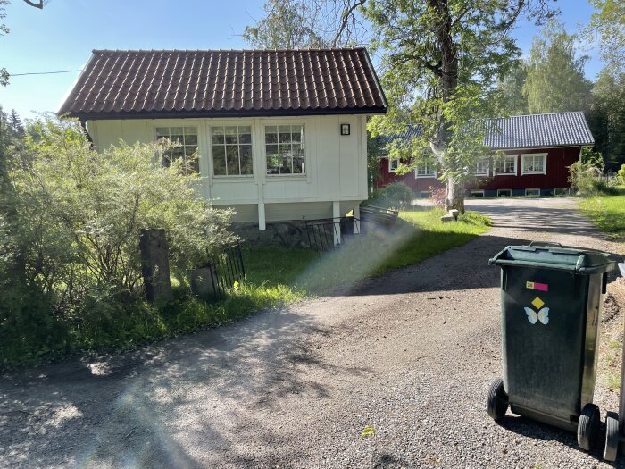 Sommardag, mindre hus, grusväg, grönska, soptunna, svensk lantlig idyll.