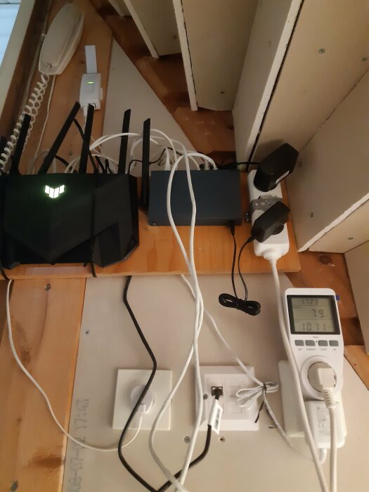 Trång teknikutrymmesvrå med nätverksutrustning, kablar, router, modem och strömförsörjningsenheter på trägolv under taklutning.