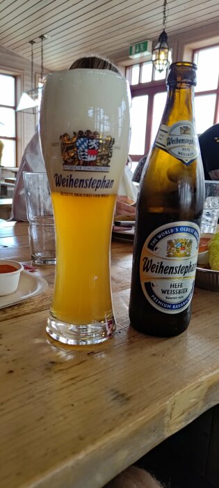 Ölglas fyllt med veteöl bredvid tom flaska, Weihenstephan, restauranginteriör i bakgrunden.