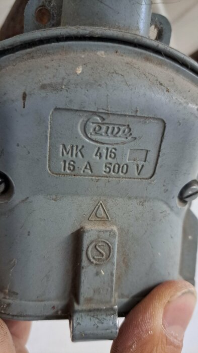 En person håller i en grå metallkomponent med etikett "MK 4,16 16.A 500V". Använd, smutsig, industriell look.
