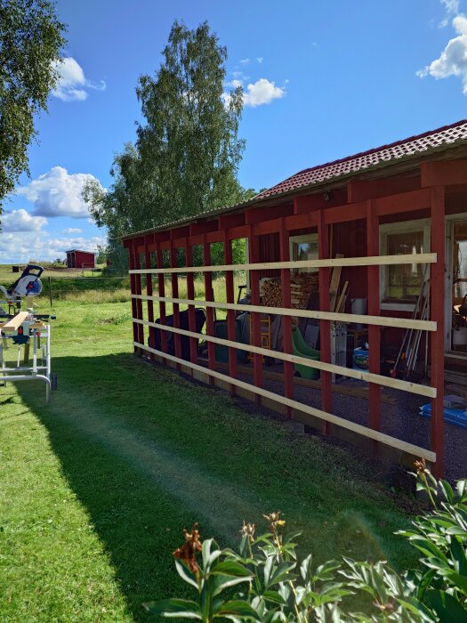 Röd stuga med veranda under konstruktion, trästapel, klar himmel, grönt gräs, landlig miljö.