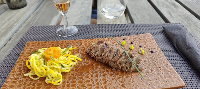 Elegant matpresentation utomhus med skivad köttbit, garnering, ett glas vin och vattenglas på träbord.