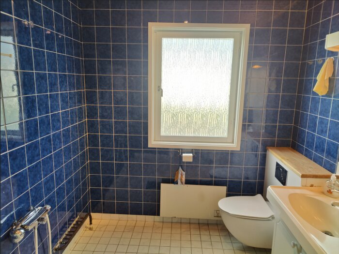 Ett badrum med blå kakel, toalett, handfat, frostad fönsterglas och handdukar.