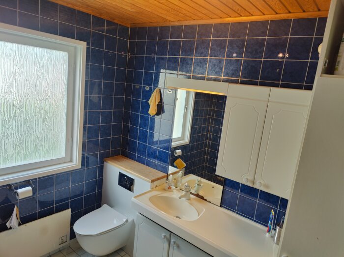 Badrum med blå kakel, trädetaljer, toalett, handfat, spegelskåp och frostat fönster.