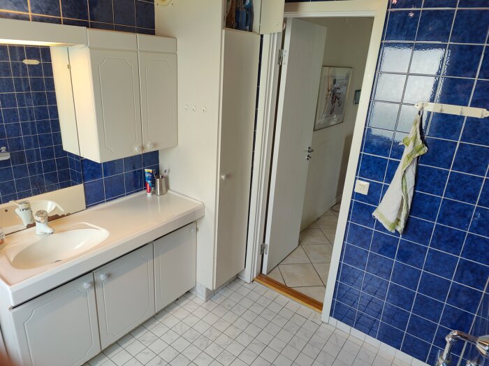 Badrum med blå kakel, vit skåp, handfat, spegel och en öppen duschdörr.