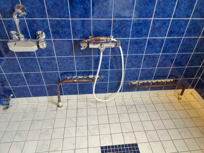 Ett duschkabin med blå kakelväggar, vit duschslang, kran, och rostiga rör syns.