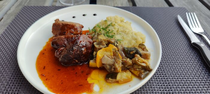 Ett tallrik med kött, sås, mos och grönsaksröra serverad på ett utomhusbord med bestick.