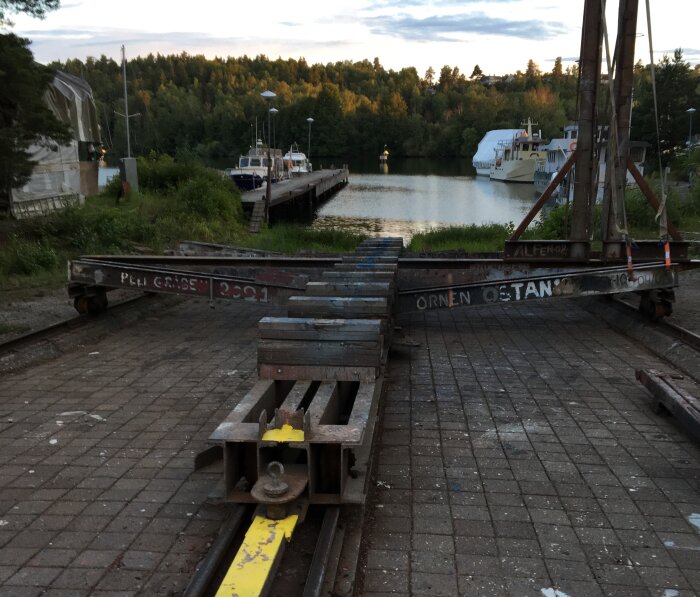 Hamn med slipvagn på räls, båtar vid brygga, träd, kvällsljus.