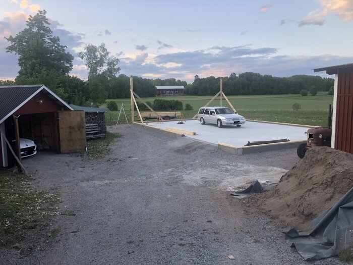 Byggplats med färdig grund, träställning, två bilar, lada, hög med jord, och landskap i skymningen.