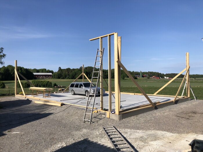 Konstruktion av trästomme, blå himmel, bil, landsbygd, gröna fält, grusväg, byggnadsställning, stege.