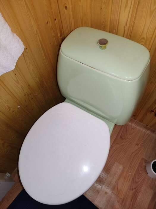 Gammaldags toalett i enkelt badrum med träväggar och parkettgolv. Vit sits, ljusgrön cistern.