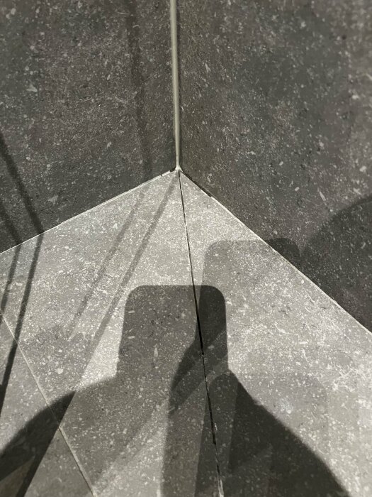 Skugga av en person på gråa stenplattor nära ett hörn. Kontrast, ljus, geometri och textur framträder.