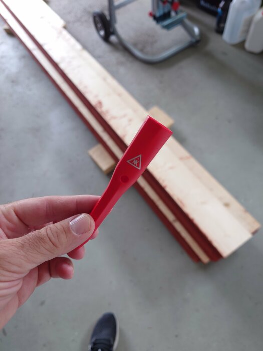Hand håller röd platt föremål i verkstad, träplankor och handtruck i bakgrunden.
