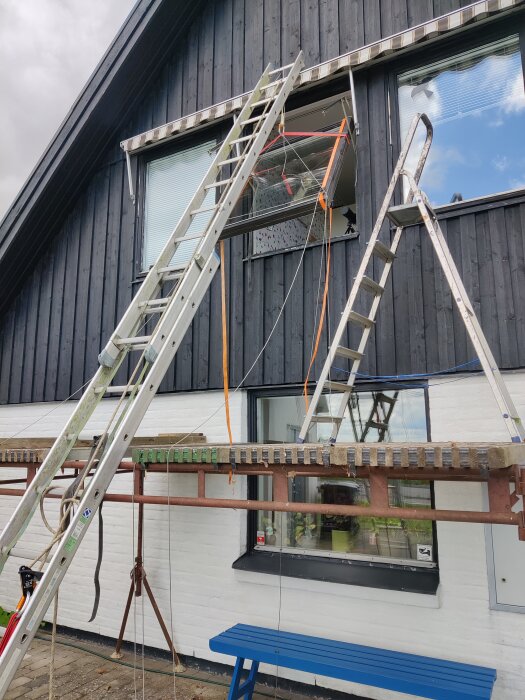 Två stegar står mot ett hus, komplicerad uppsättning med stag och rep för säkerhet eller arbete på höjd.