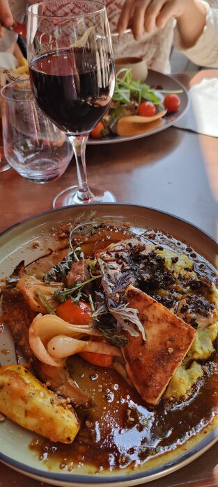 Måltid med pasta, grönsaker, tomater. Ett glas rödvin framför. Person i bakgrunden. Ljust, trevligt inslag.