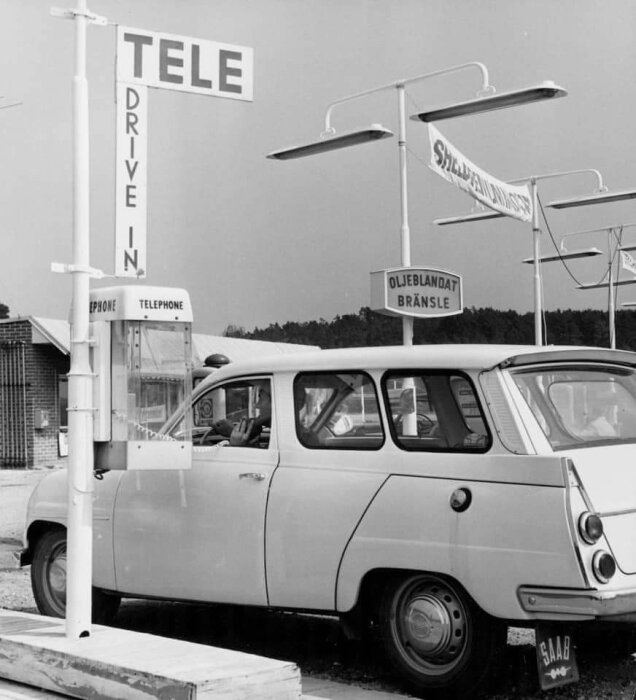 Svartvit bild: äldre bil vid drive-in telefonstation, retro, skyltar med "TELE", "SHELL", "Oljeblandat bränsle".